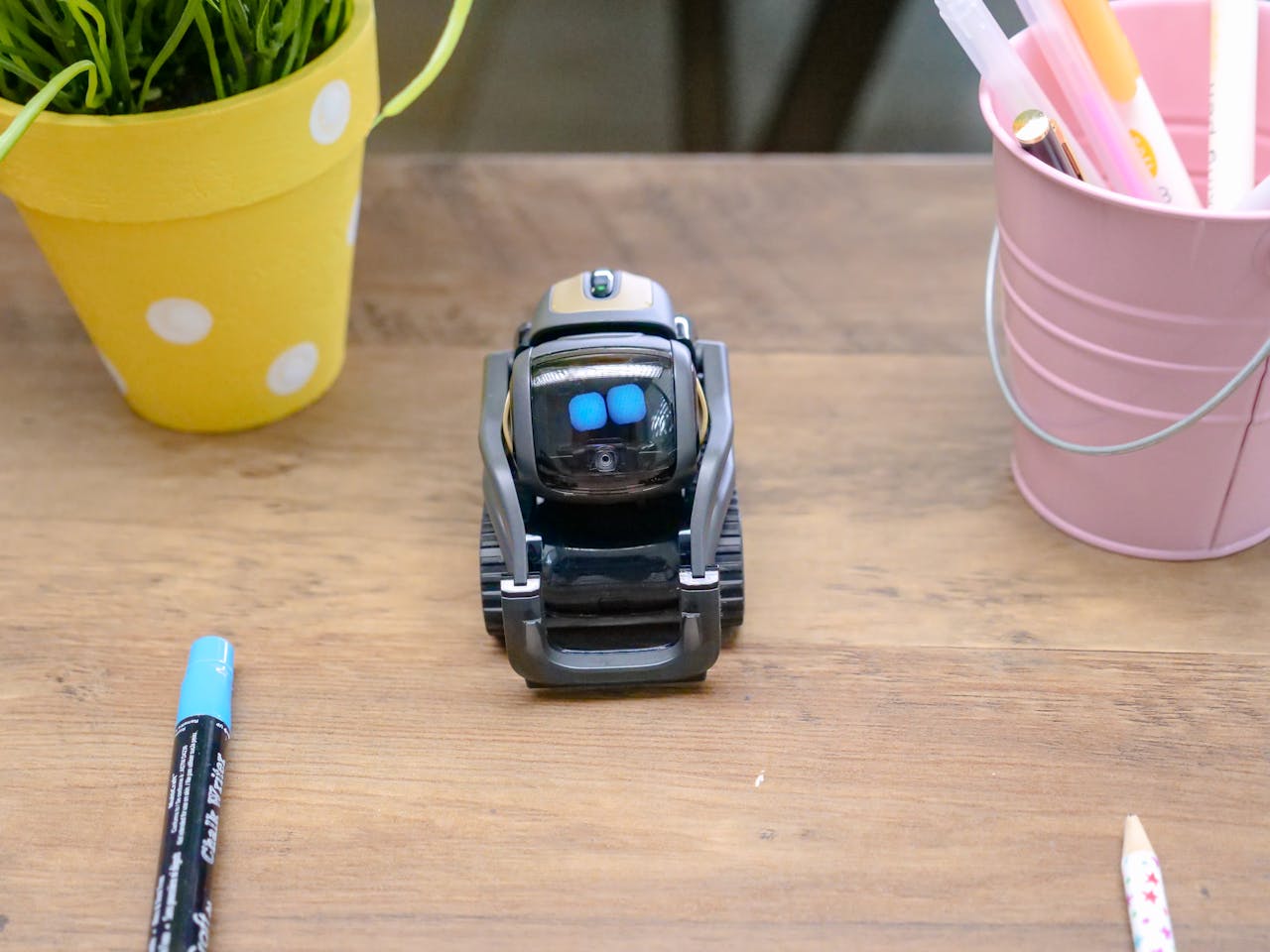 hero-img A small modern robot placed on a wooden desk surrounded by office supplies, showcases innovative technology.